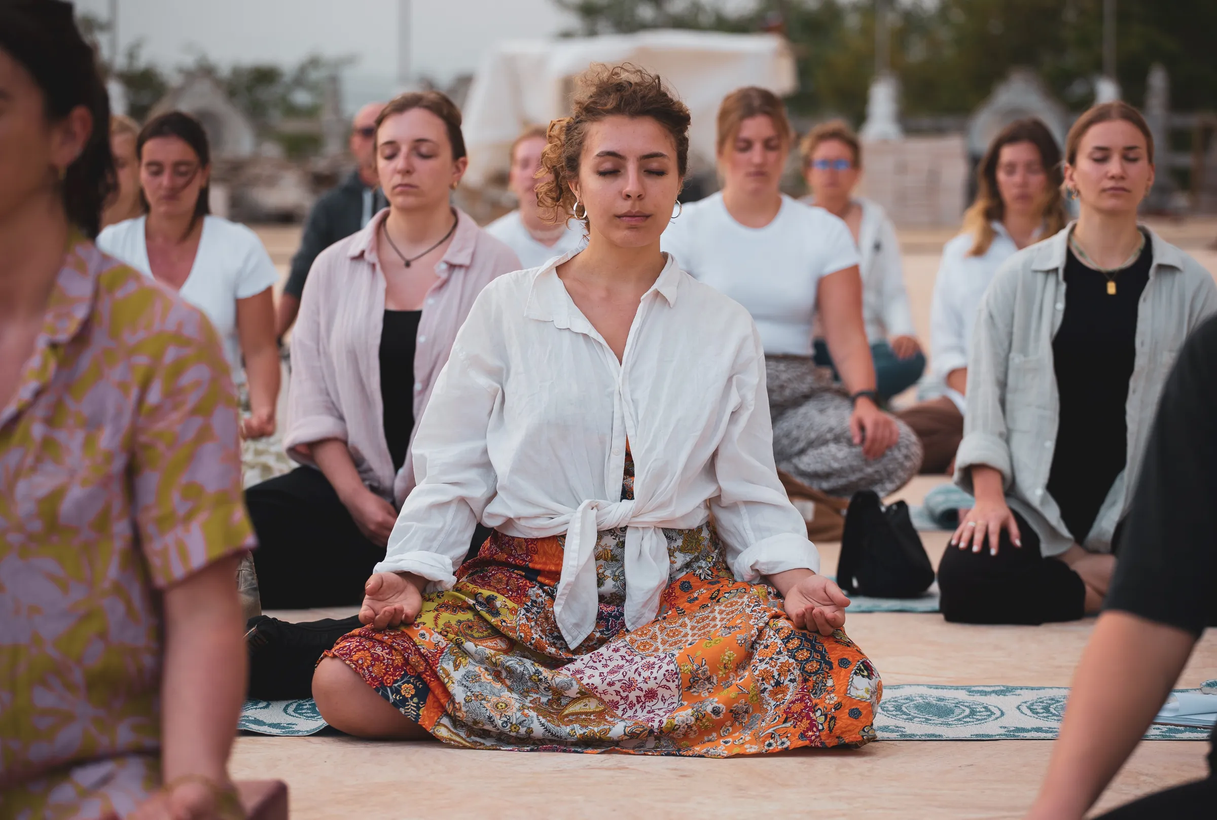 A group of people sitting cross-legged outdoors, meditating together with their eyes closed at dusk.