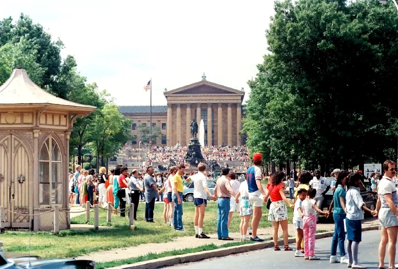 A long human chain of people holding hands along a tree-lined path, with a crowd gathered before a grand neoclassical building.